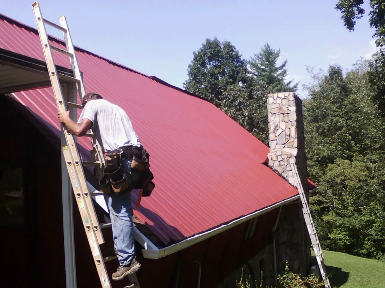 Roofer on a ladder beside a red metal roof and stone chimney — Soffit & Fascia Repair by Roofing Brothers in Leesport